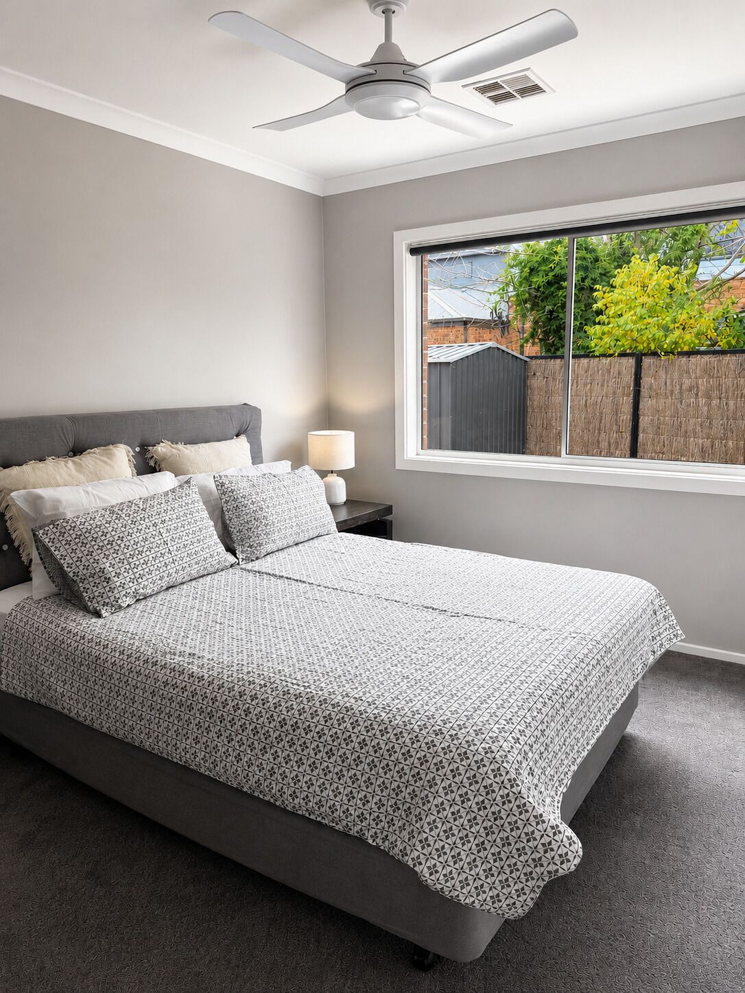 A bedroom with a bed, a nightstand, a lamp, a ceiling fan, and a window with a view of a backyard.