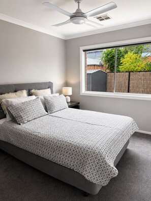 A bedroom with a bed, a nightstand, a lamp, a ceiling fan, and a window with a view of a backyard. - Comfort on Clarke (Moama)