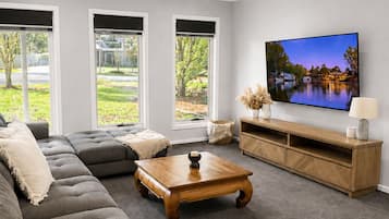 A modern living room with a sectional sofa, a wooden coffee table, a TV, and a large window with a view of greenery.