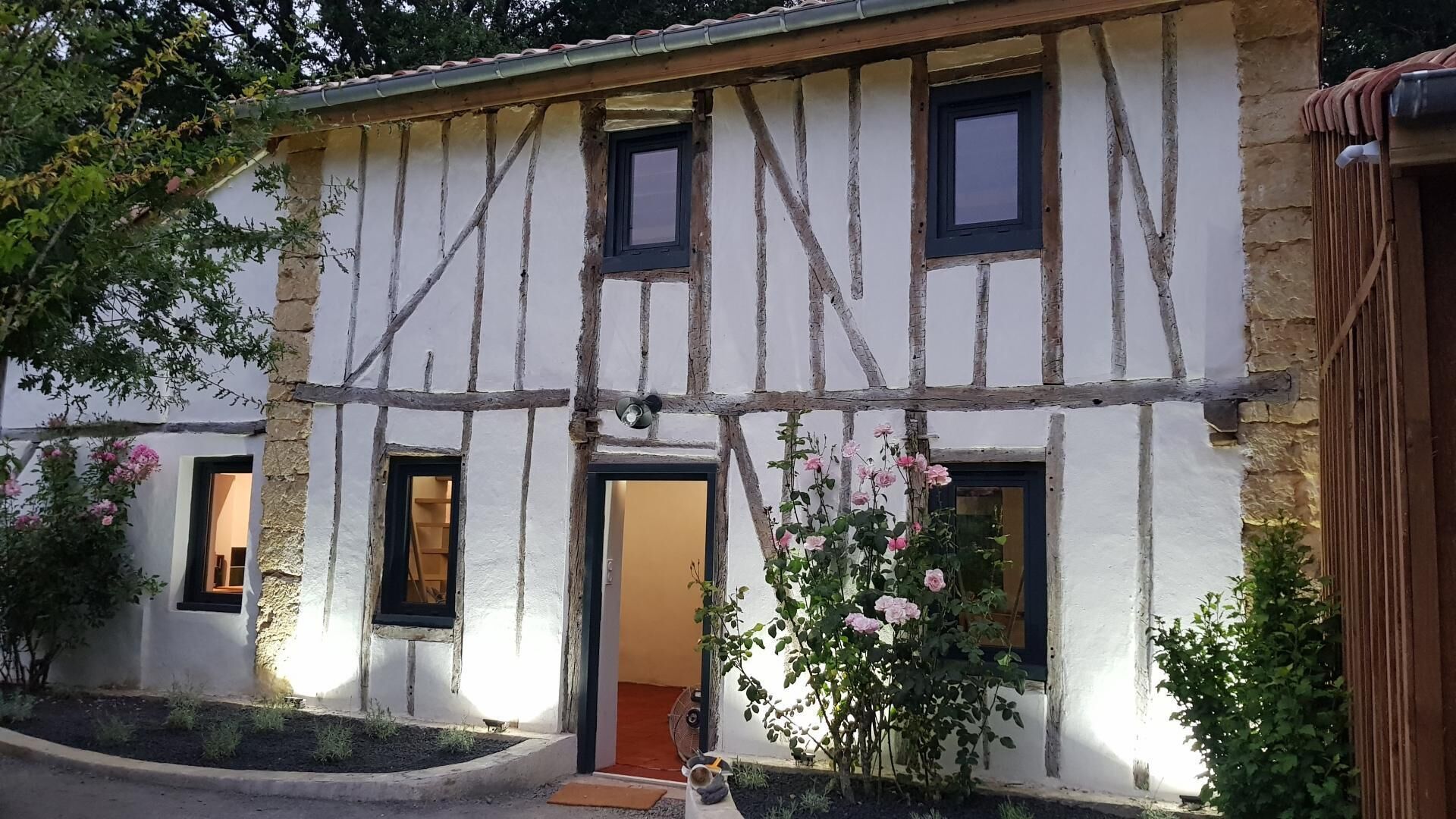 A half-timbered house with two visible windows, a door, and a small garden with pink flowers.