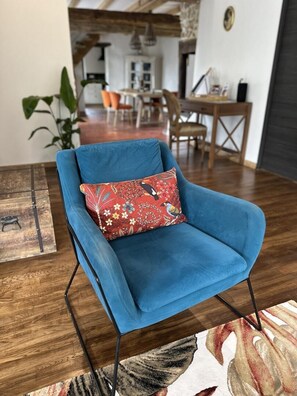 A blue armchair with a floral pillow, positioned on a patterned rug in a room with wooden flooring and a dining area in the background. - My Country House (EUGENIE LES BAINS)