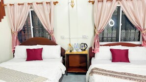 Habitación de hotel con dos camas, cada una con almohada y manta, una mesita de noche con hervidor y botellas de agua, y cortinas.
