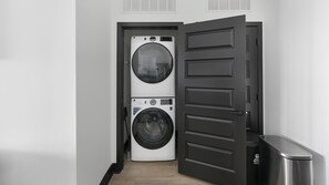 A laundry room with a front-loading washer and dryer, a trash can, and a door with a handle. - Landing | Stunning 1BD (Nashville)