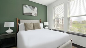 Une chambre d’hôtel avec un grand lit, deux tables de chevet avec des lampes, un mur vert et une fenêtre avec des stores.