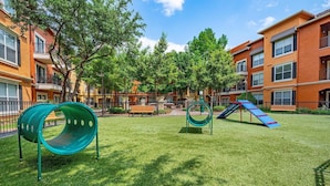 Een speeltuin met glijbanen, waaronder een groene spiraalvormige glijbaan, omringd door een verzorgd gazon en bomen, met appartementen op de achtergrond.