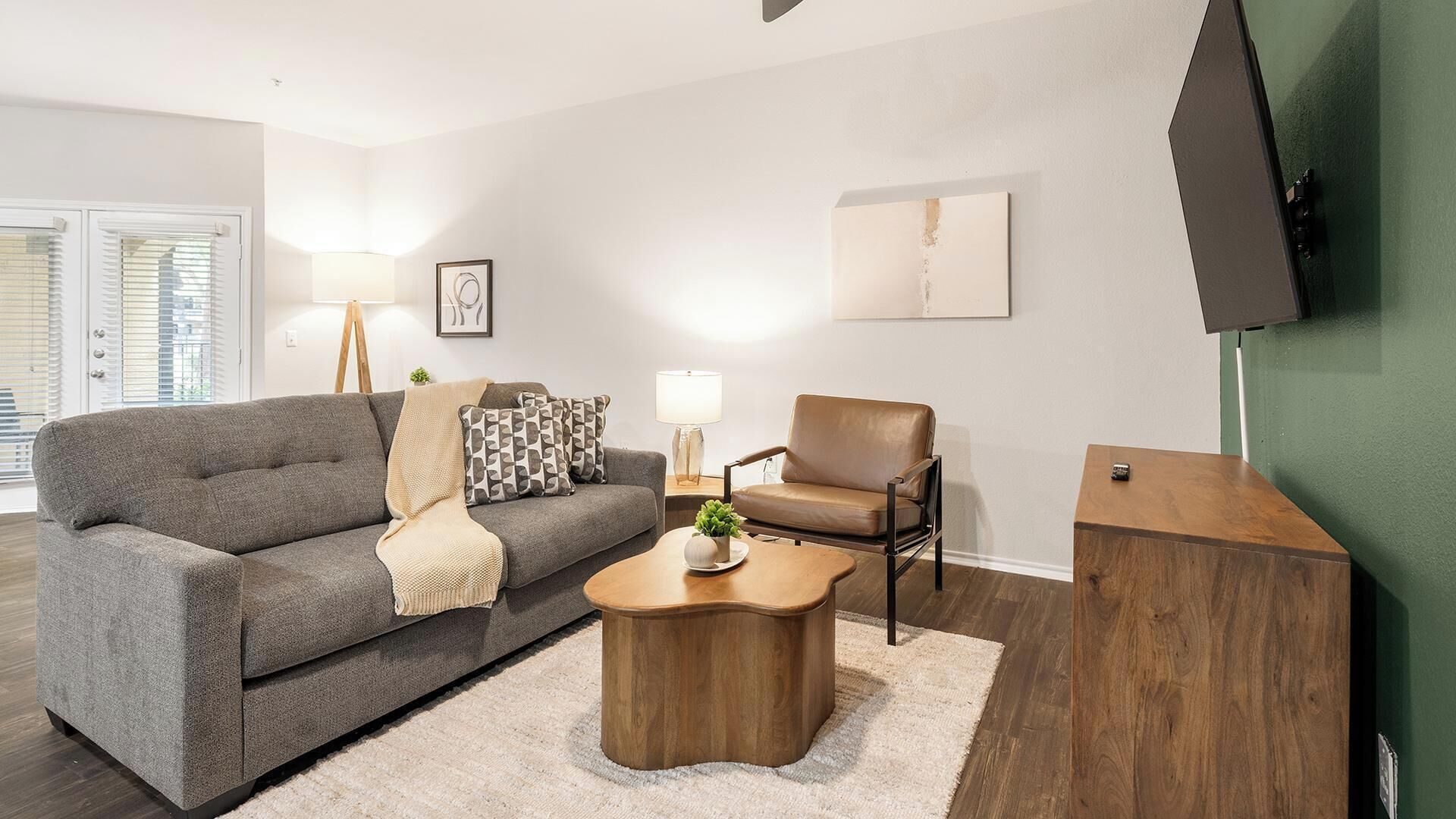 Un salon avec un canapé gris, un fauteuil en cuir marron, une table basse en bois et un téléviseur fixé au mur.