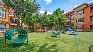 Een speeltuin met glijbanen, waaronder een groene spiraalvormige glijbaan, omringd door een verzorgd gazon en bomen, met appartementen op de achtergrond.