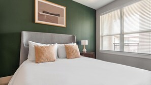 Quarto de hotel com cama, mesa de cabeceira, luminária e janela com persianas.