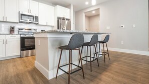 A modern kitchen with white cabinetry, stainless steel appliances, and a granite countertop. There are three bar stools with metal frames and cushioned seats. - Landing | Beautiful 2BD, Courtyard, Gym (San Antonio)