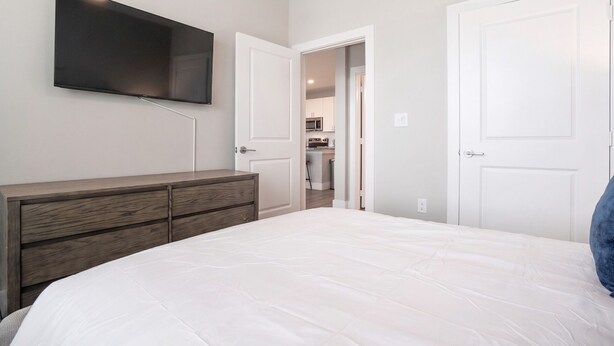 A bedroom with a bed, a dresser, a television mounted on the wall, and a door leading to a kitchenette. - Landing | Stunning 2BD, Gym, Courtyard (San Antonio)