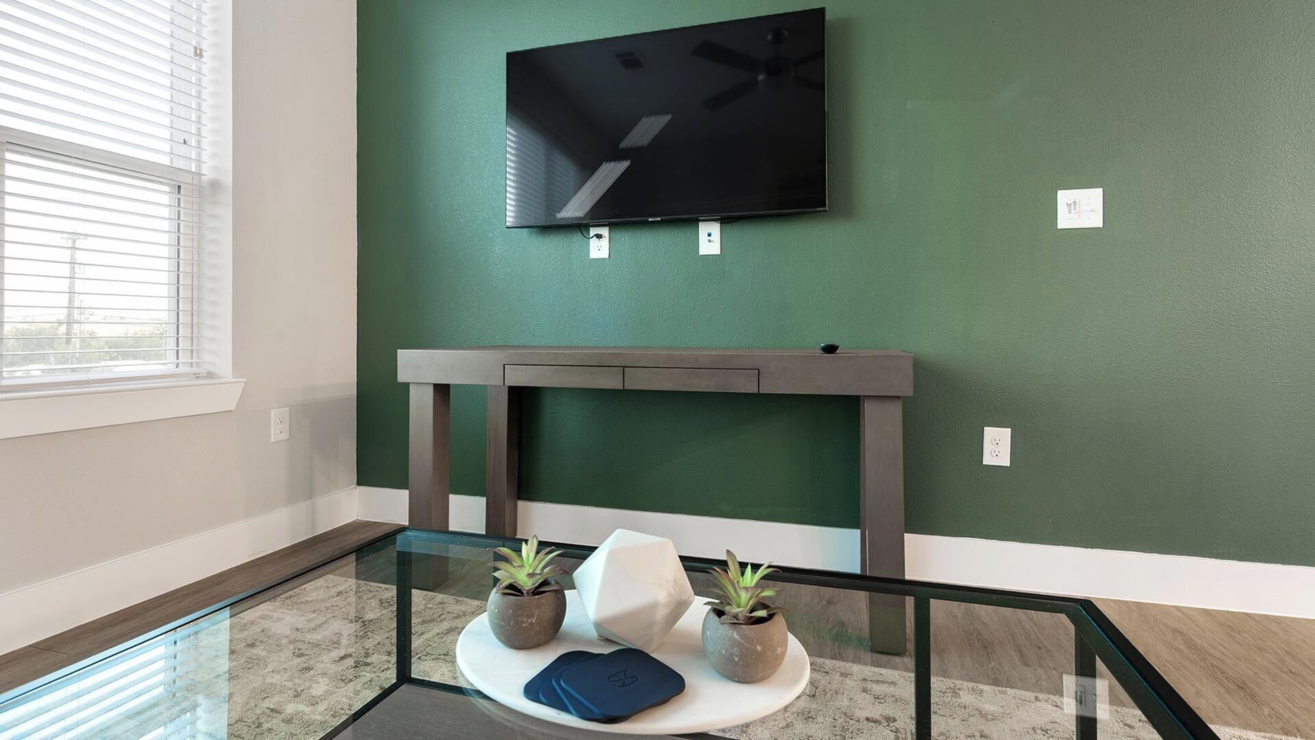 A room with a glass-top table, two potted plants, a white cushion, and a flat-screen TV mounted on a green wall.