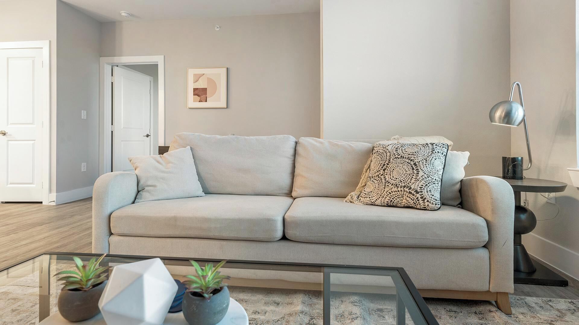 A neutral-toned living room with a sofa, a glass coffee table, and two potted plants.
