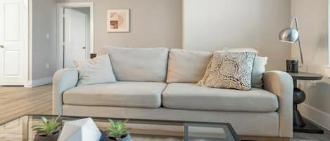 A neutral-toned living room with a sofa, a glass coffee table, and two potted plants.