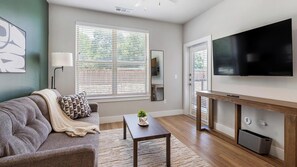 A modern living room with a sofa, a coffee table, a TV, and a large window with blinds. - Landing | Modern 1BD, Clubhouse, Gym (Fort Worth)