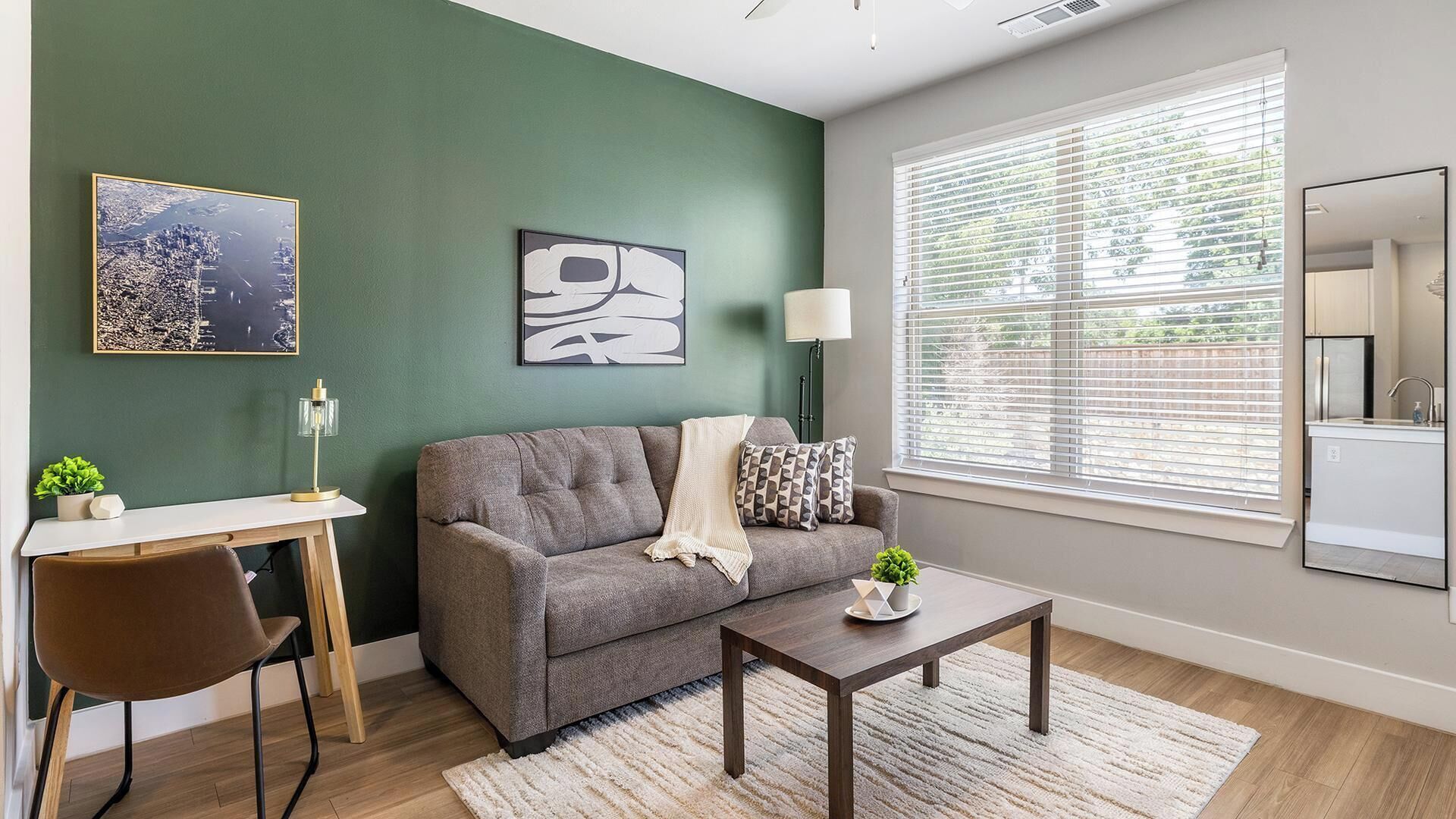 A living room with a grey sofa, a coffee table, a desk with a chair, a lamp, and artwork on the walls.