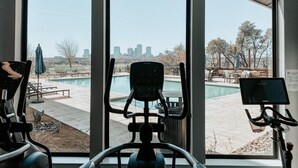 A gym with exercise equipment and a view of a swimming pool and city skyline. - Landing | Modern 1BD, Gym, Clubhouse (Fort Worth)