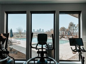 Ein Fitnessraum mit Trainingsgeräten und Blick auf ein Schwimmbecken sowie Stadtpanorama.