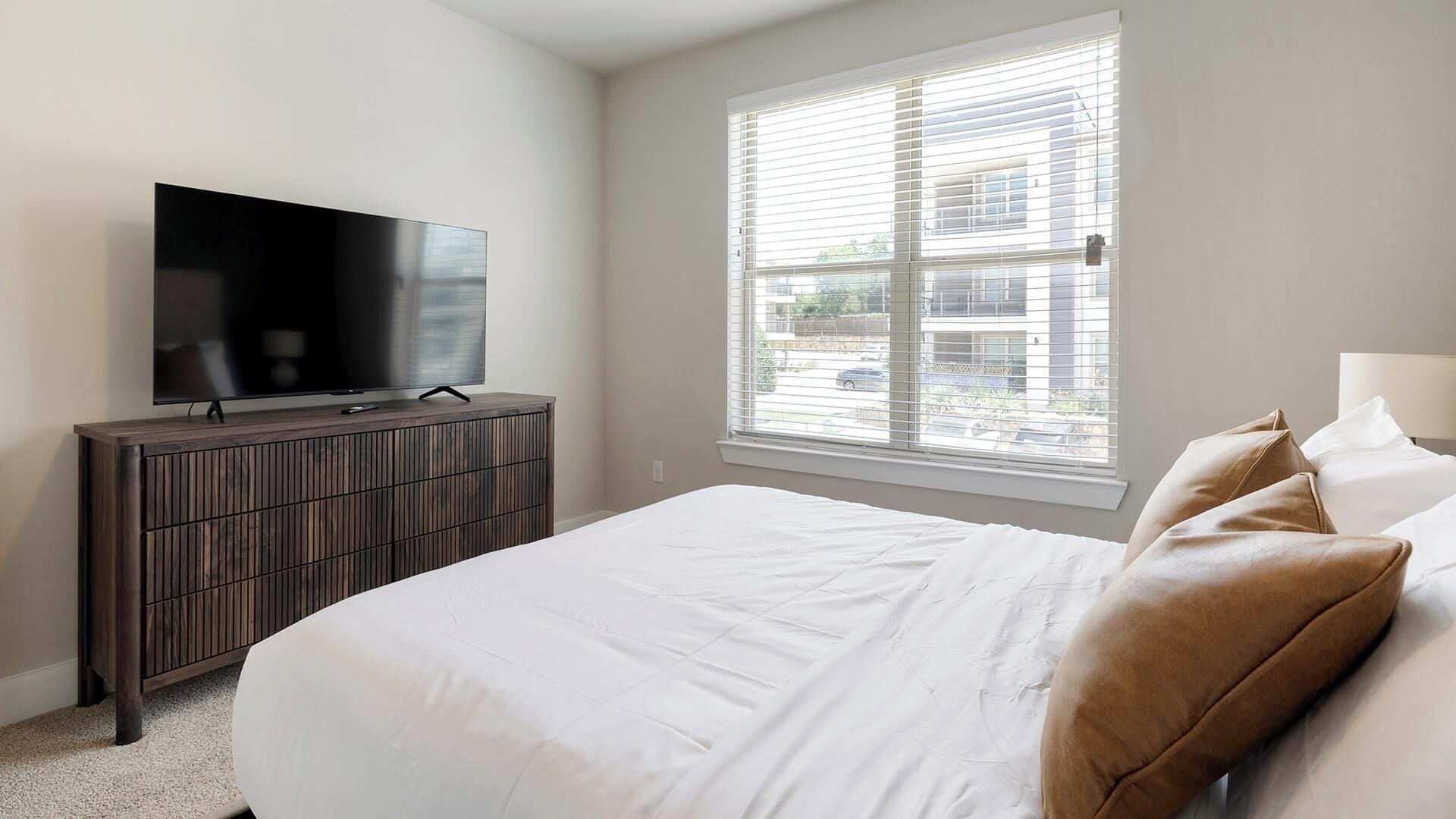 A bedroom with a bed, a television on a wooden dresser, and a window with blinds.