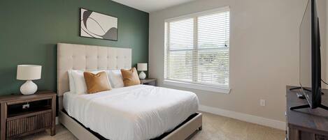 A bedroom with a large bed, a ceiling fan, a window with blinds, and a television on a stand.