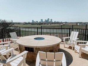 A patio with a stone fire pit, white Adirondack chairs, and a view of a city skyline. - Landing | Chic 2BD, Clubhouse, Gym (Fort Worth)