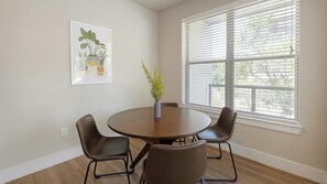 Ein Zimmer mit einem runden Tisch, vier Stühlen, einer Vase mit Pflanze, einem gerahmten Bild und einem Fenster mit Jalousien.