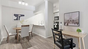 A modern dining area with a wooden table and chairs, a kitchen area with appliances, and a framed picture on the wall. - Landing | Chic 2BD, Gym, Courtyard (Allen)