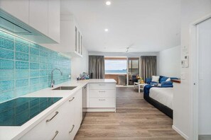 A compact hotel room with a kitchenette, a bed, a small table, and a view of the sea. - Seaview Studio Apartment (Whitianga)