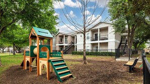Un parque infantil con tobogán y estructura para escalar, rodeado de árboles y un edificio con balcones.