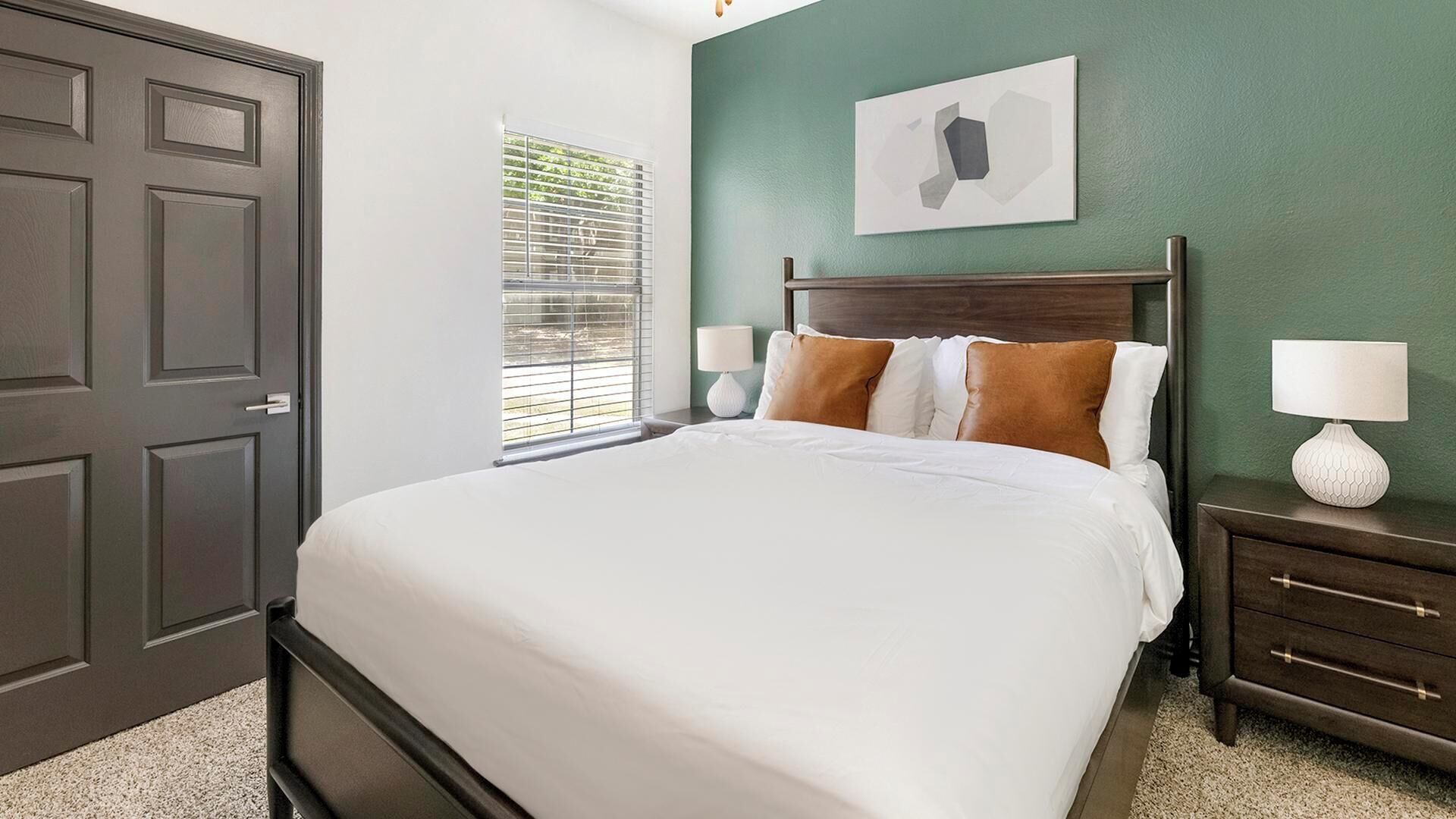 Une chambre à coucher avec un lit, une table de chevet, une lampe, une fenêtre avec des stores et une porte.