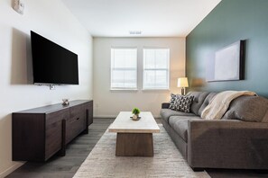 Ein modernes Wohnzimmer mit Flachbildfernseher, einer dunklen Holzkommode, einem grauen Sofa, einem hellen Couchtisch und einem Fenster mit Jalousien.