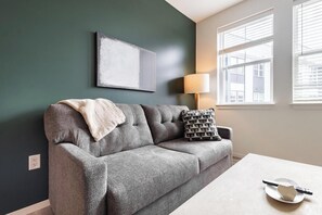 A grey sofa with a throw blanket and patterned pillows, a table with a remote and a book, a lamp, and a painting on the wall. - Landing | Spectacular 2BD in Steelpointe Harbor (Bridgeport)