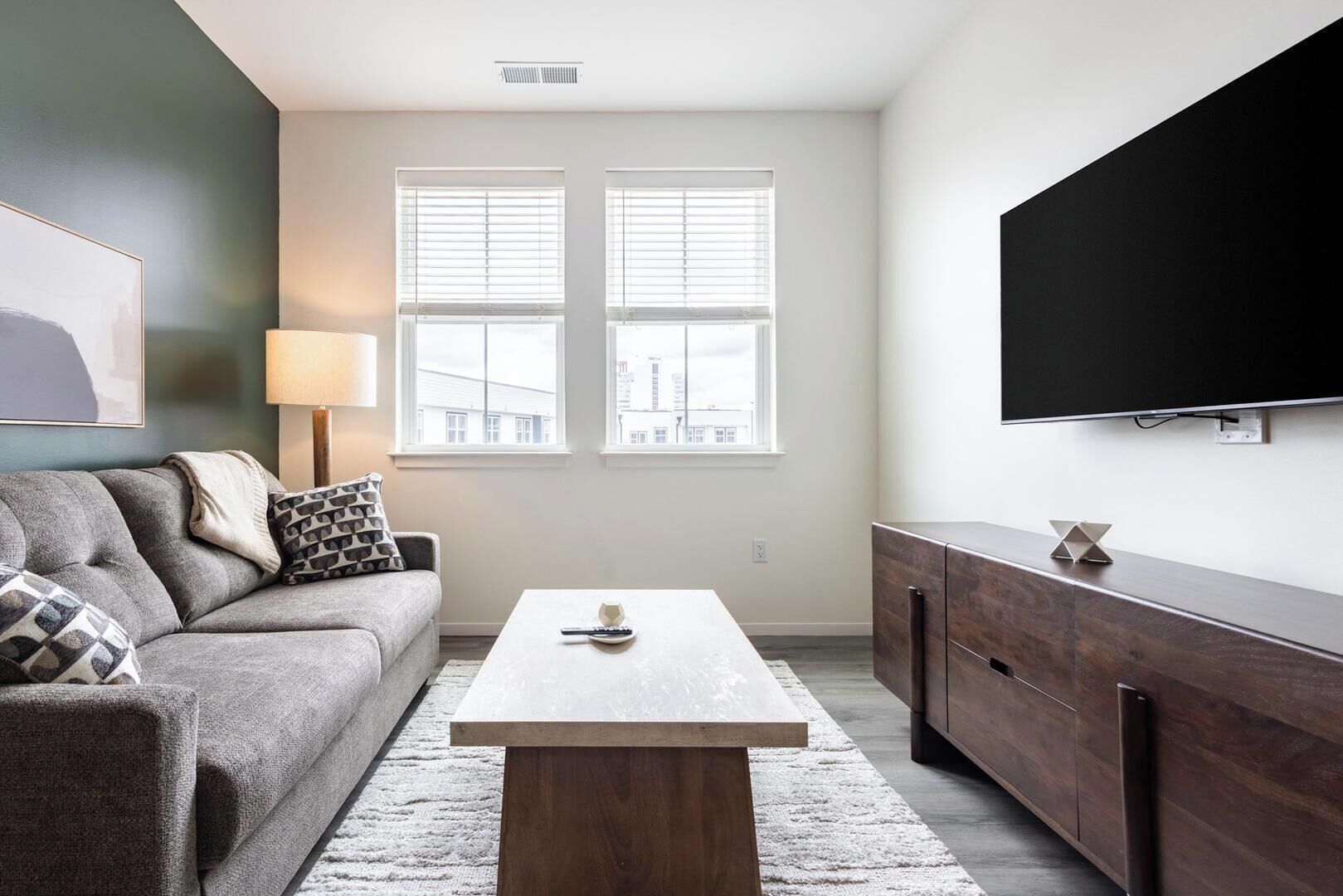 A modern living room with a grey sofa, a coffee table, a TV mounted on the wall, and a window with blinds.