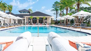 Une piscine entourée de chaises longues et d’un parasol, bordée de palmiers et d’un bâtiment doté de grandes fenêtres.