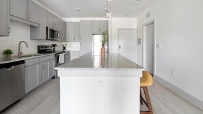 A modern kitchen with grey cabinets, a stainless steel dishwasher, a microwave, and a central island with a bar stool. - Landing | Chic 1BD, Gym (St. Augustine)