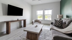 A living room with a flat-screen TV mounted on the wall, a wooden coffee table, a grey sofa with patterned cushions, and a window with blinds. - Landing | Chic 1BD, Gym (St. Augustine)