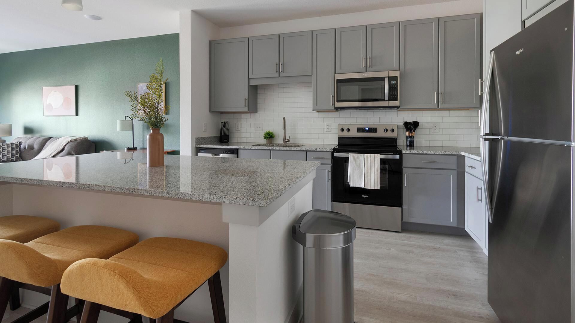 Une cuisine moderne avec des meubles gris, un réfrigérateur en acier inoxydable, un four à micro-ondes et une cuisinière. On y trouve également un coin bar avec des tabourets et une poubelle.