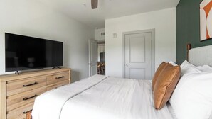 Un dormitorio con cama, televisor sobre una cómoda de madera y puerta que da a un baño.