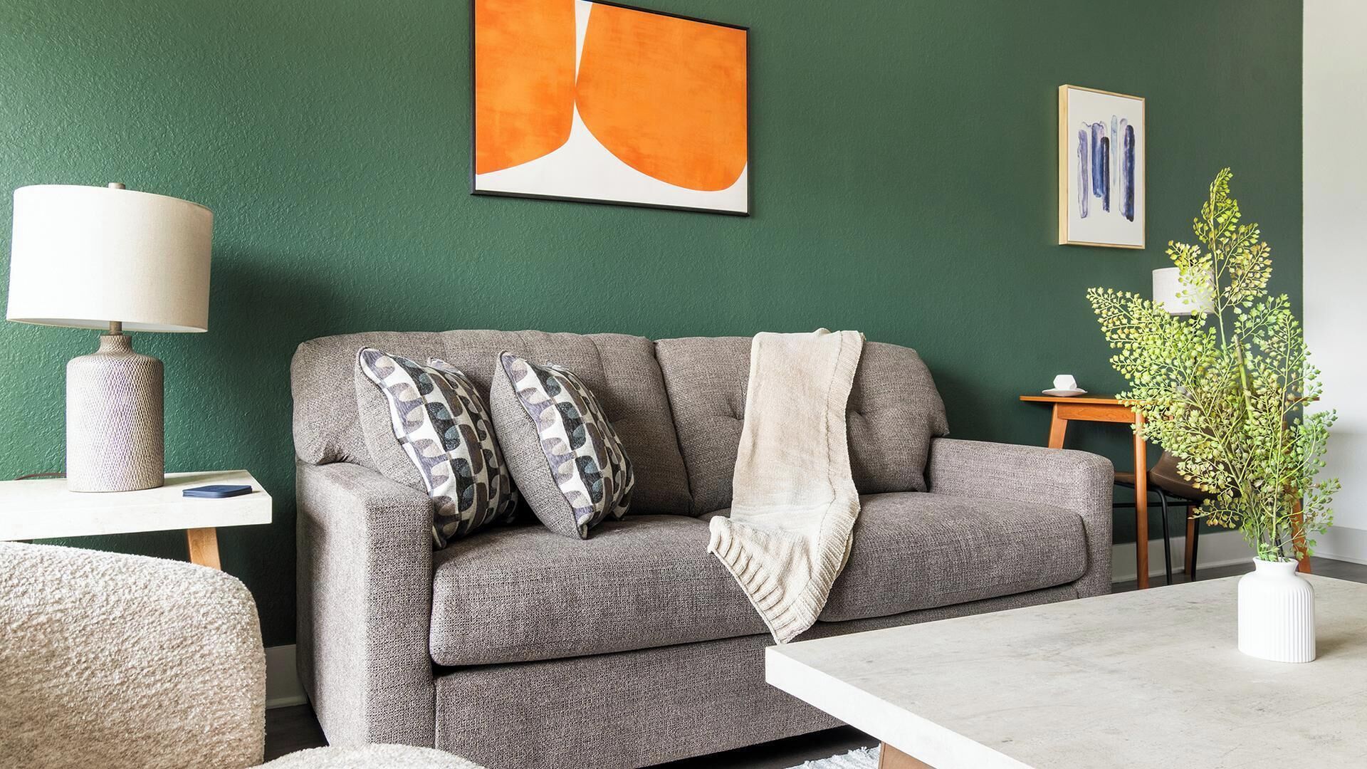 Een woonkamer met een grijze bank, kussens met een patroon, een witte plaid, een lamp en een vaas met een plant.
