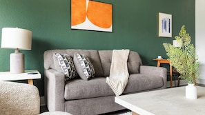 Een woonkamer met een grijze bank, kussens met een patroon, een witte plaid, een lamp en een vaas met een plant.