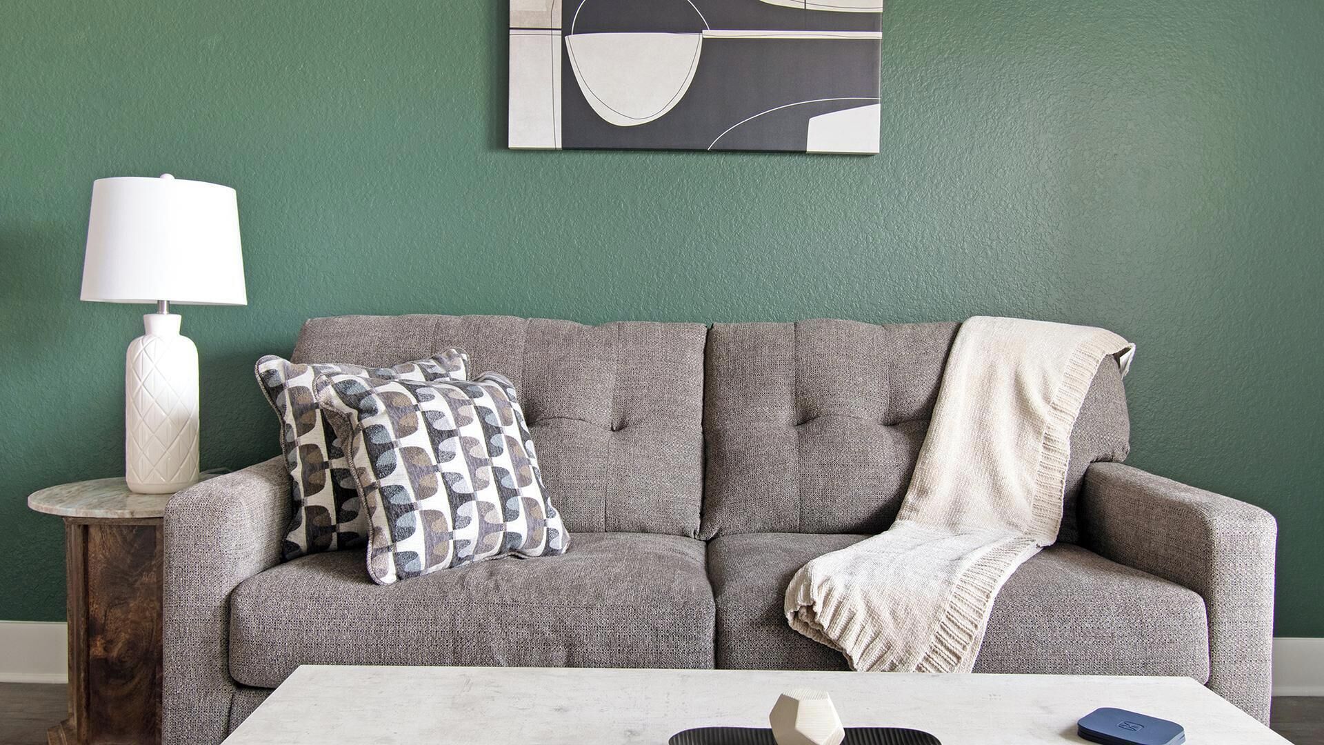 A grey sofa with a patterned pillow and a throw blanket, a white table with a lamp and a book, and a green wall with a geometric artwork.