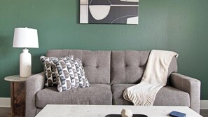 A grey sofa with a patterned pillow and a throw blanket, a white table with a lamp and a book, and a green wall with a geometric artwork. - Landing | Stylish 2BD, Gym (St. Augustine)