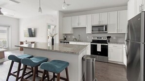 A modern kitchen with white cabinets, a stainless steel refrigerator, a microwave, and an oven. There are three bar stools with teal upholstery. - Landing | Incredible 2BD, Gym (St. Augustine)