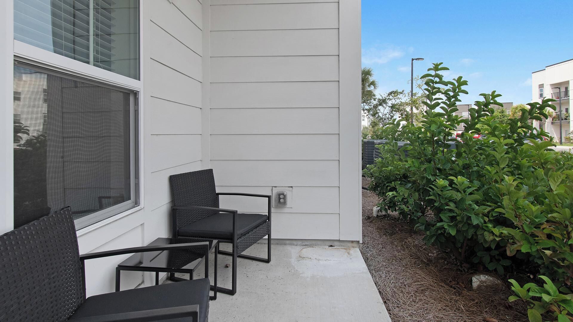 A patio with two chairs and a table, a white exterior wall, and a small garden area with green shrubs.