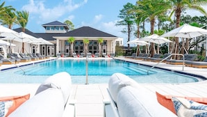Une piscine entourée de chaises longues et d’un parasol, bordée de palmiers et d’un bâtiment doté de grandes fenêtres.