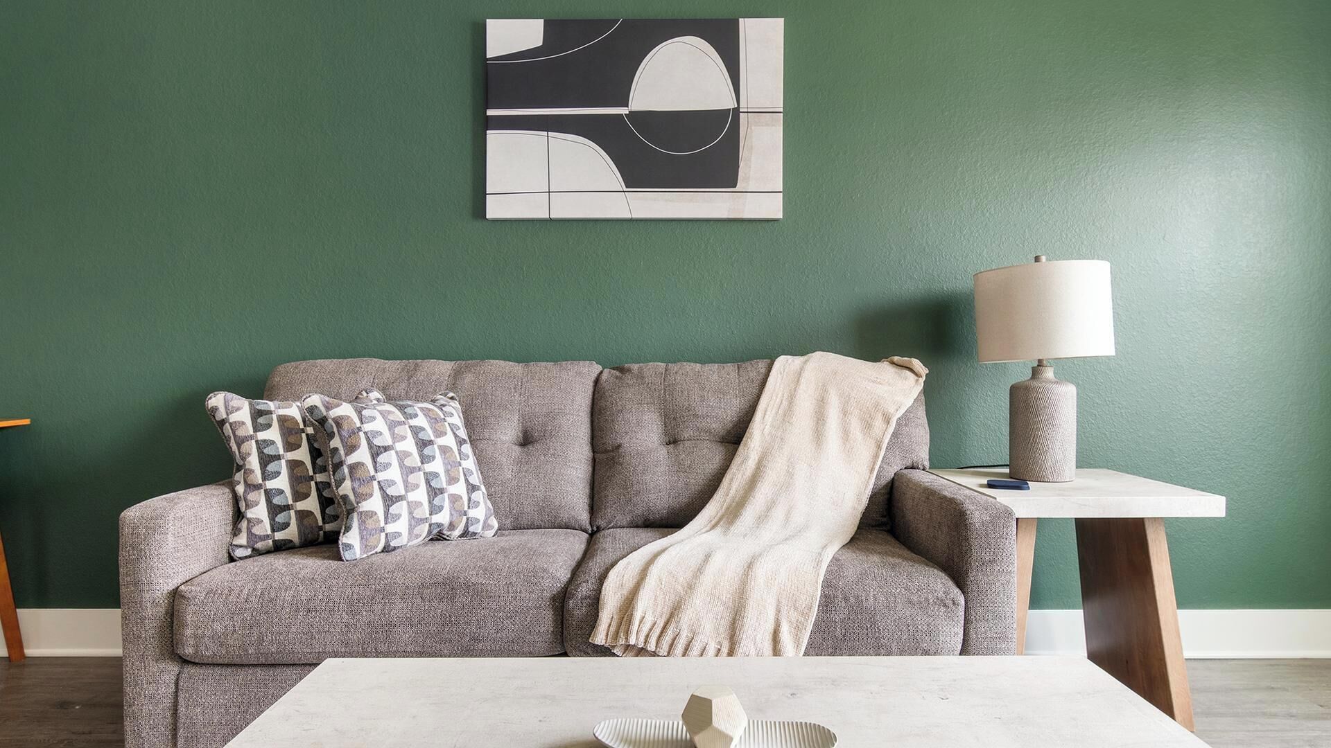 A living room with a grey sofa, patterned pillows, a white throw blanket, a white coffee table, and a wall-mounted lamp.