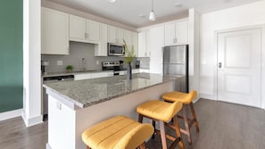 A modern kitchen with white cabinets, a granite countertop, stainless steel appliances, and two yellow bar stools. - Landing | Beautiful 1BD, Gym (St. Augustine)