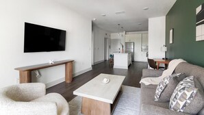 A modern living room with a flat-screen TV, a wooden bench, a white coffee table, a grey sofa with patterned cushions, and a kitchen in the background. - Landing | Spectacular 1BD, Gym (St. Augustine)