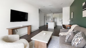 Ein modernes Wohnzimmer mit einem Flachbildfernseher, einer hölzernen Bank, einem weißen Couchtisch, einem grauen Sofa mit gemusterten Kissen und einer Küche im Hintergrund.