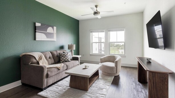 A living room with a green wall, a sofa, a coffee table, a chair, a TV, and a ceiling fan. - Landing | Spectacular 1BD, Gym (St. Augustine)