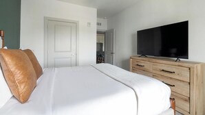 A bedroom with a bed, a television on a wooden dresser, and a door leading to a bathroom. - Landing | Spectacular 1BD, Gym (St. Augustine)